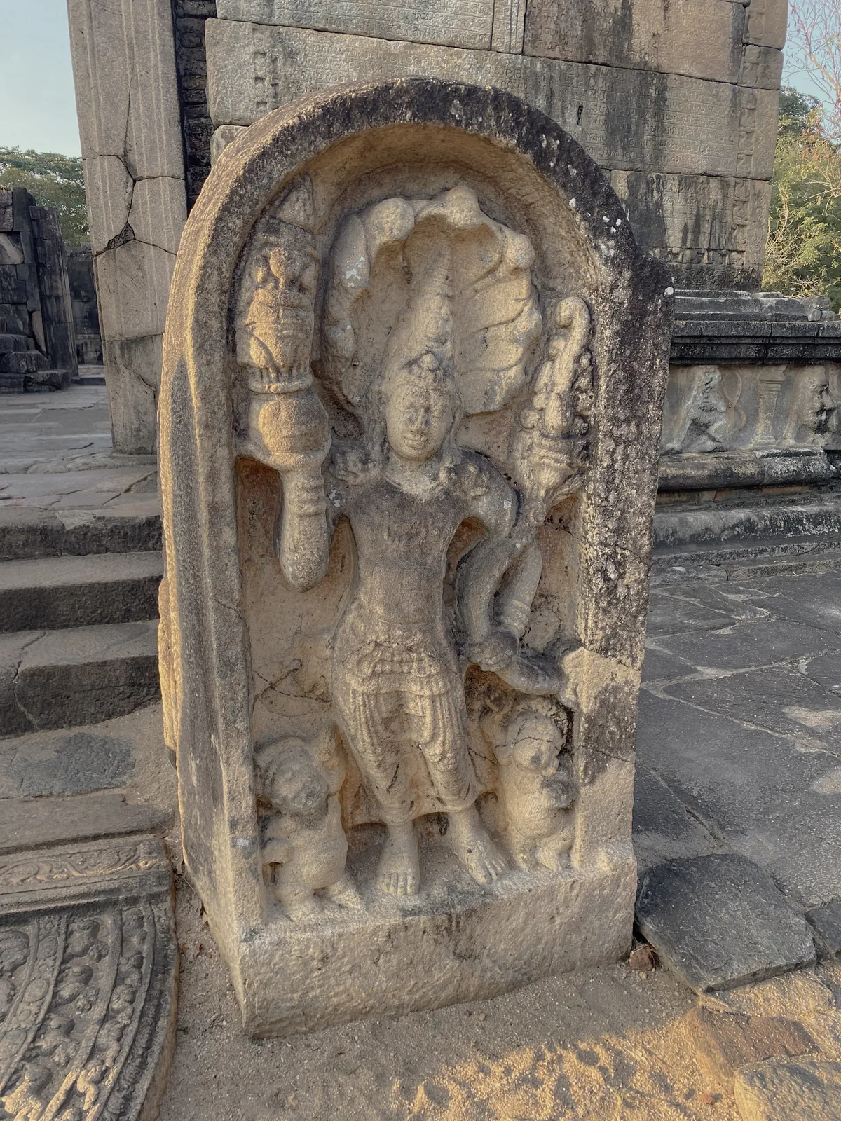 A Nagaraja guardstone at the Polonnaruwa Vatadage, a crowned serpent-king holding a pot of plenty beneath a cobra hood, a dwarf at his feet.