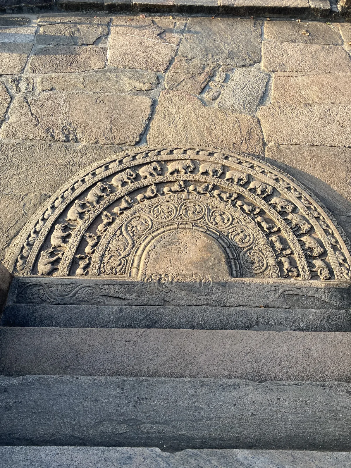 The moonstone at the Polonnaruwa Vatadage, a semicircular stone carved in fine concentric bands of animals, creeper and a central half-lotus, seen at an angle.