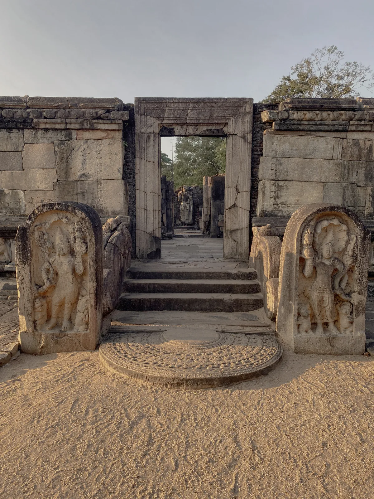 An entrance stair of the Polonnaruwa Vatadage, a moonstone at the foot, a carved Nagaraja guardstone at each side, and a seated Buddha visible through the doorway.